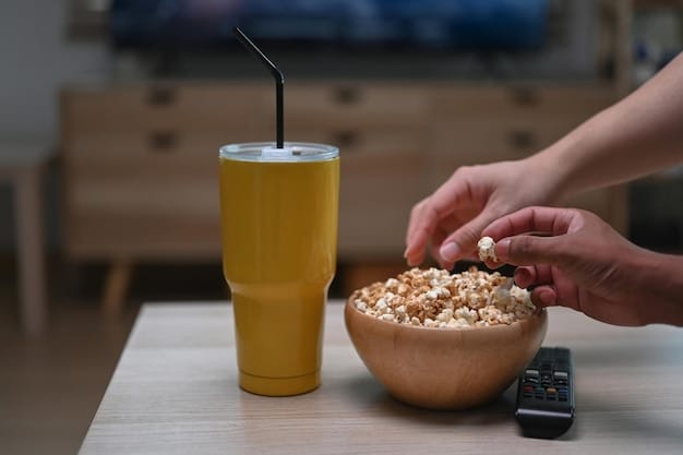A close-up of hands holding a bowl of popcorn and a bottle of water, resting on a soft blanket during a cozy binge-watching session. The TV screen is blurred in the background, focusing on the comfort essentials.