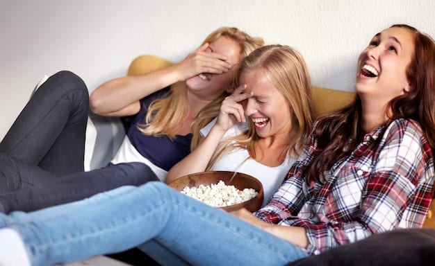 A group of friends laughing and sharing popcorn on a comfortable couch, all looking towards a television screen that is out of frame. The room is dimly lit, suggesting a relaxed, evening viewing session.