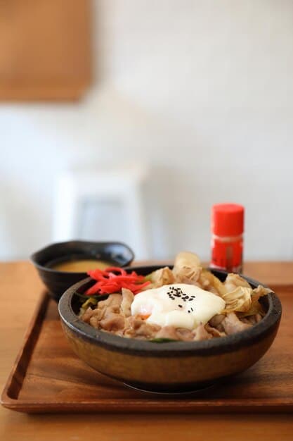 A close-up shot of a steaming bowl of Budae Jjigae (Army Stew) with ramen noodles, slices of spam, sausages, kimchi, and tofu, garnished with spring onions, captured on a rustic wooden table.